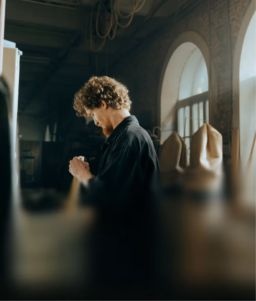 Man with curly hair and beard working with hands in a dimly lit workshop with arched windows.
