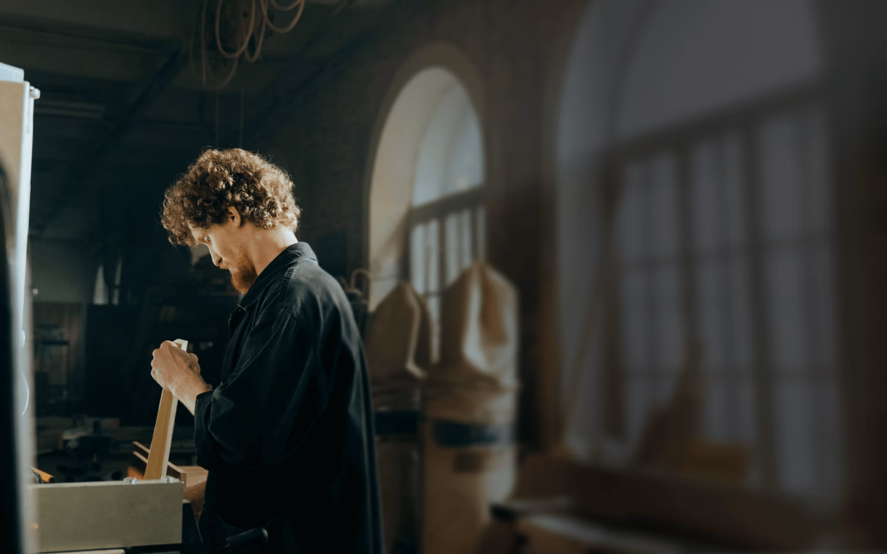 Man with curly hair and beard working with a wooden plank in an architecture workshop with arched windows.