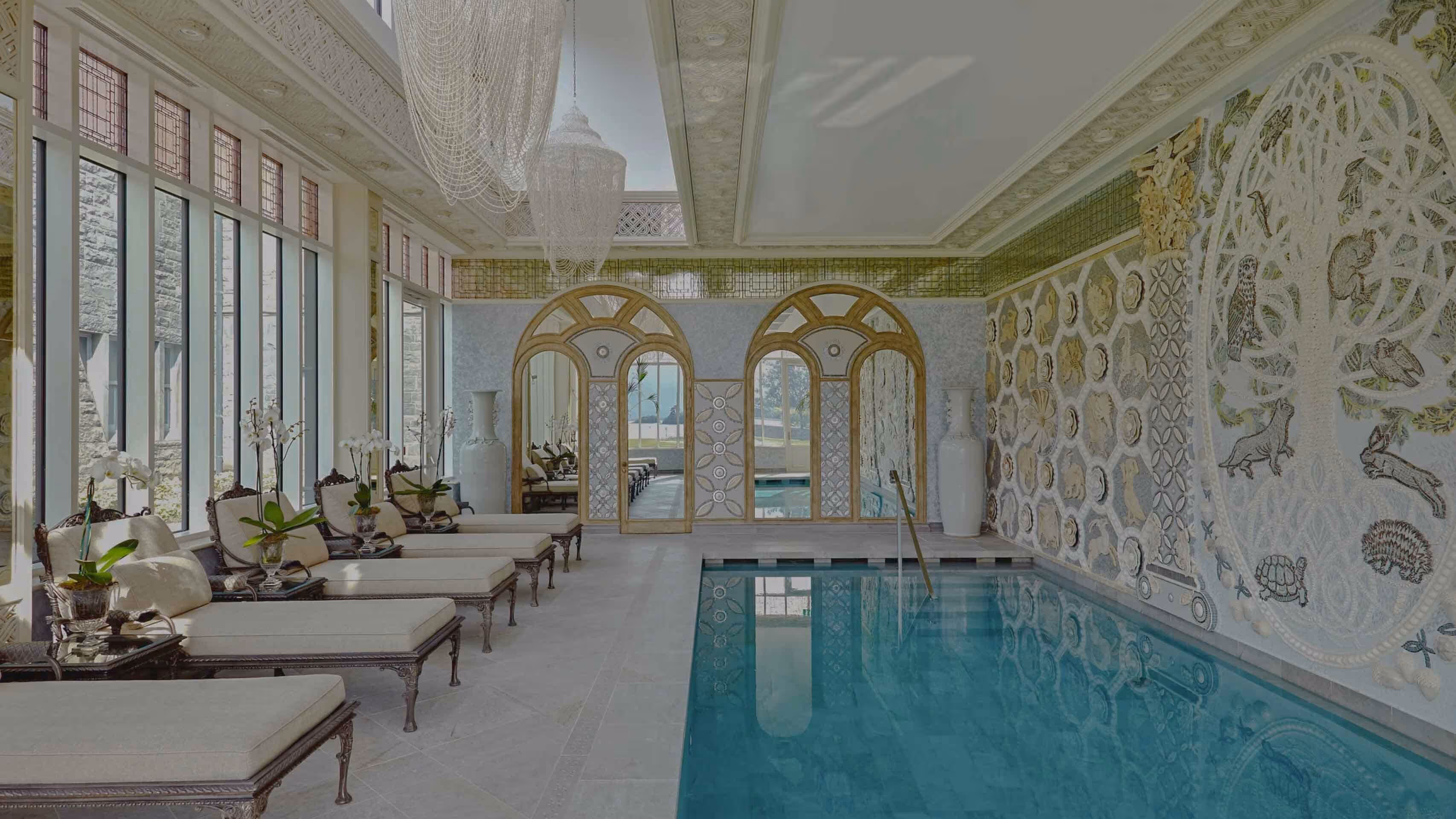 Elegant indoor pool area with lounge chairs, large windows, ornate ceiling, and nature-themed mosaic wall art.